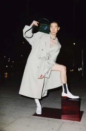 Man wearing a light grey trench coat and white boots posing outdoors at night holding a dark green handbag.