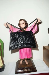 Statue of child figure in pink robe holding a large black quilted handbag with chain straps
