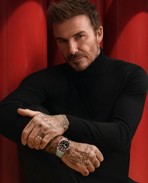 Close-up portrait of David Beckham in a black turtleneck, featuring arm tattoos and a watch, set against a plain red backdrop.