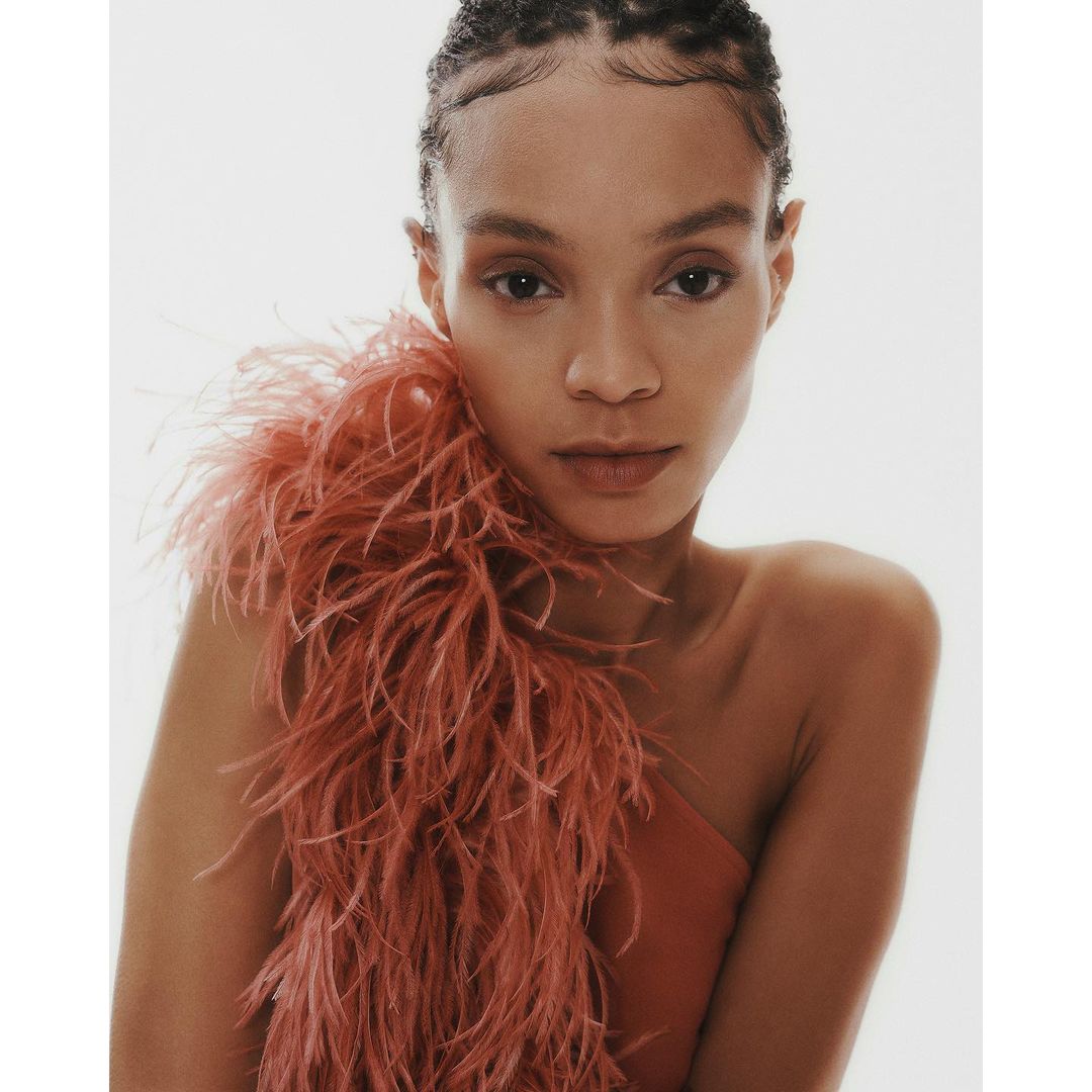 Close-up portrait of a woman with braided hair wearing a coral one-shoulder top and feathered accessory against a white background.