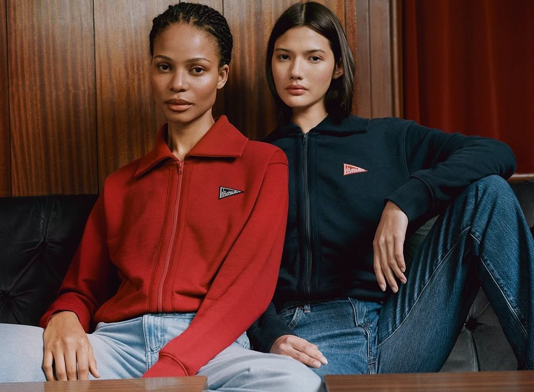 Two fashion models pose indoors, wearing matching zip-up jackets and jeans in a medium shot, with a wood-panelled wall backdrop.