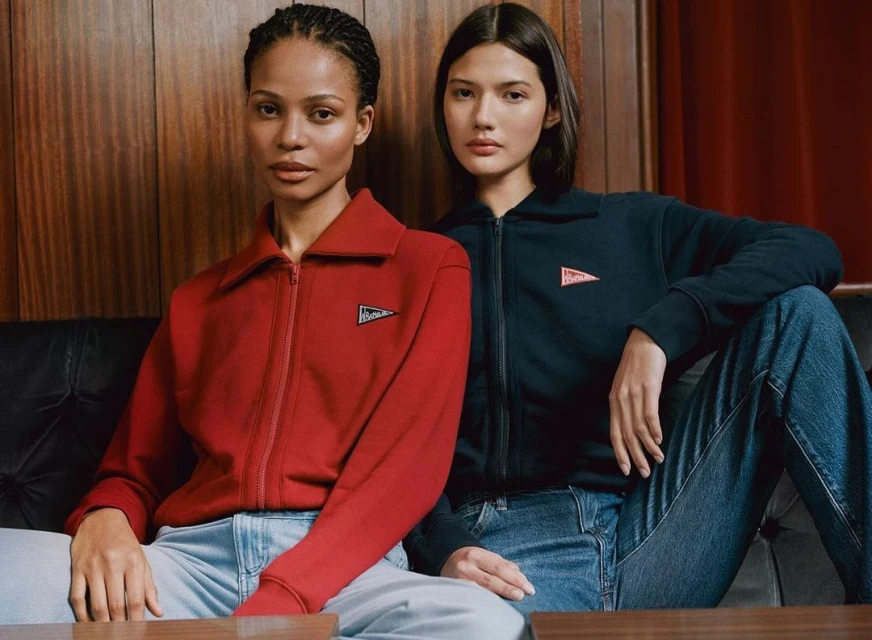 Two fashion models pose indoors, wearing matching zip-up jackets and jeans in a medium shot, with a wood-panelled wall backdrop.