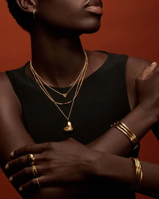 Close-up shot of an African American female model showcasing gold jewelry: layered necklaces, rings, and multiple bangle bracelets against a red background.