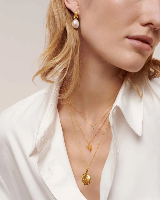 Close-up of a fair-skinned woman wearing gold pendant necklaces and pearl earrings, adorned in a white blouse, studio setting.