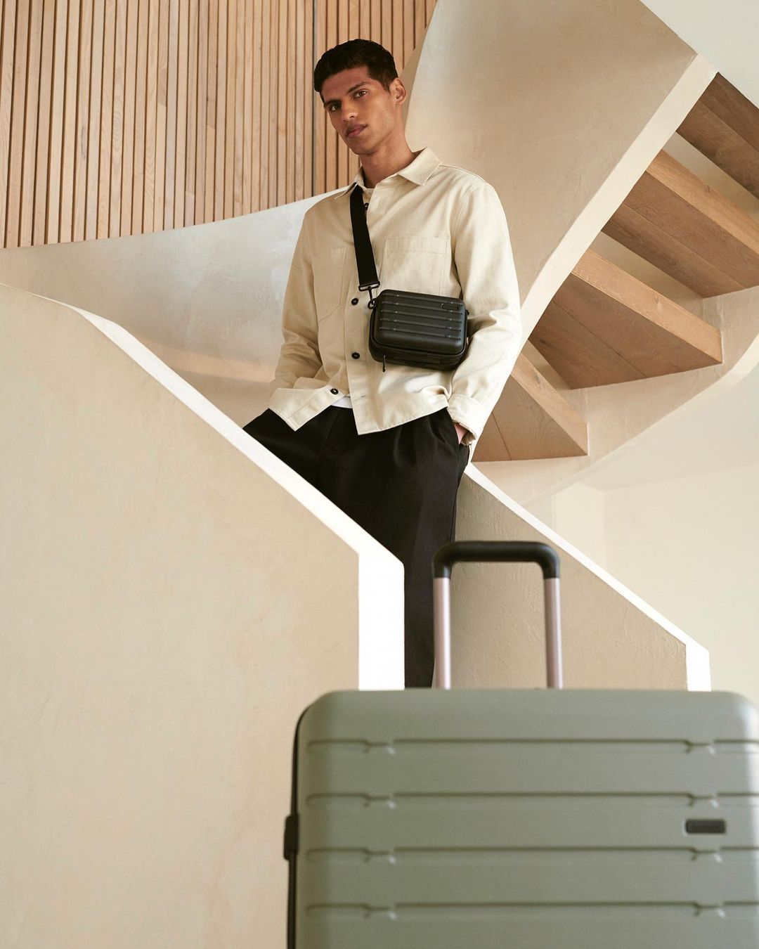Man on stairs with black crossbody bag and olive green rolling suitcase. Neutral tones and modern design enhance the scene.