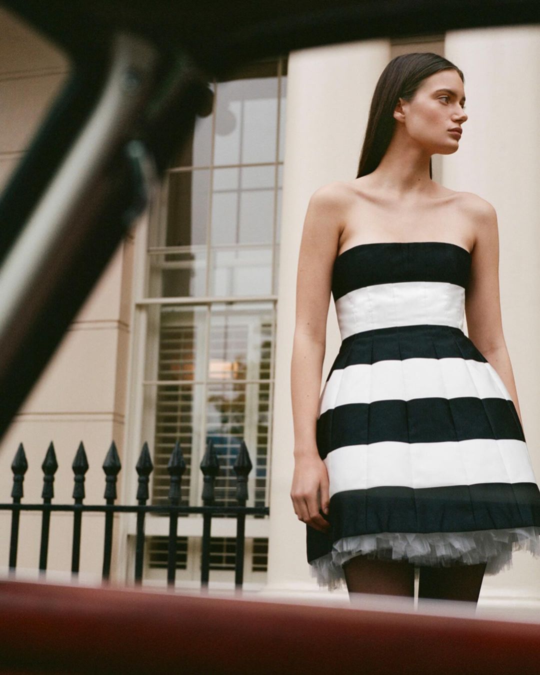 An adult female poses in a black and white strapless dress in front of a beige building with a black metal fence, captured outdoors.