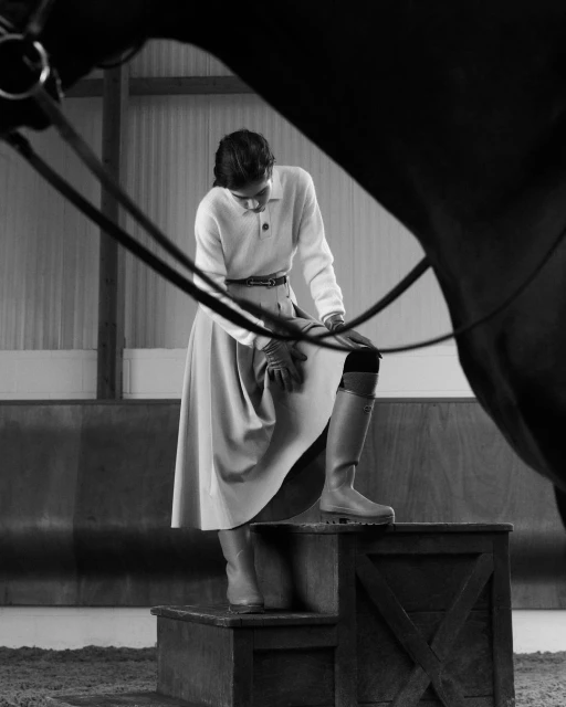 Woman wearing riding boots and skirt standing on a wooden platform next to a horse in an indoor equestrian setting.