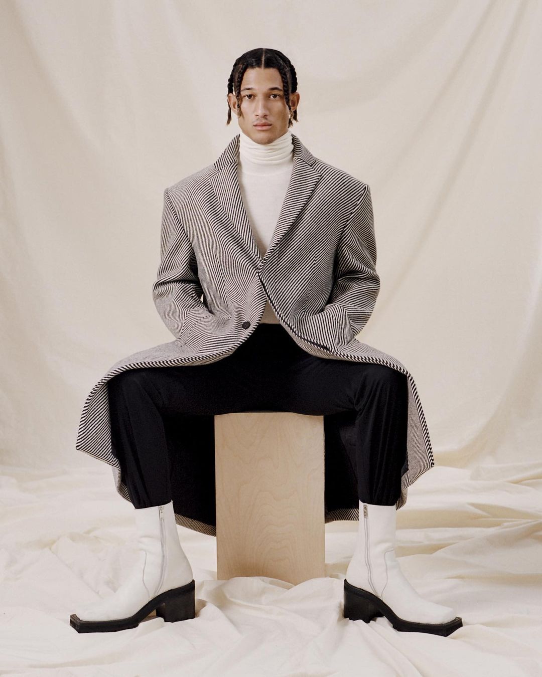 Portrait of a Black male model sitting on a wooden block in a checkered coat, white turtleneck, black pants, and white boots in a studio.