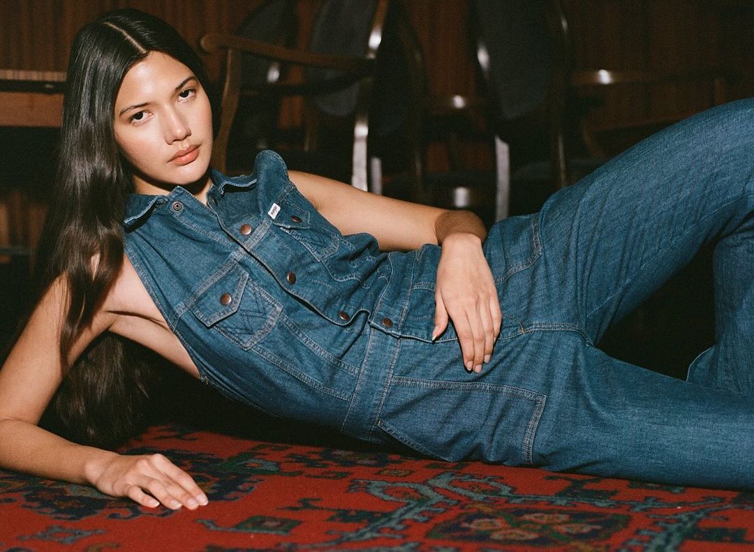 Woman in blue denim jumpsuit lying on a red patterned carpet. She's indoors with hardwood furniture in the background.