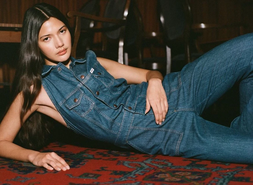 Woman in blue denim jumpsuit lying on a red patterned carpet. She's indoors with hardwood furniture in the background.