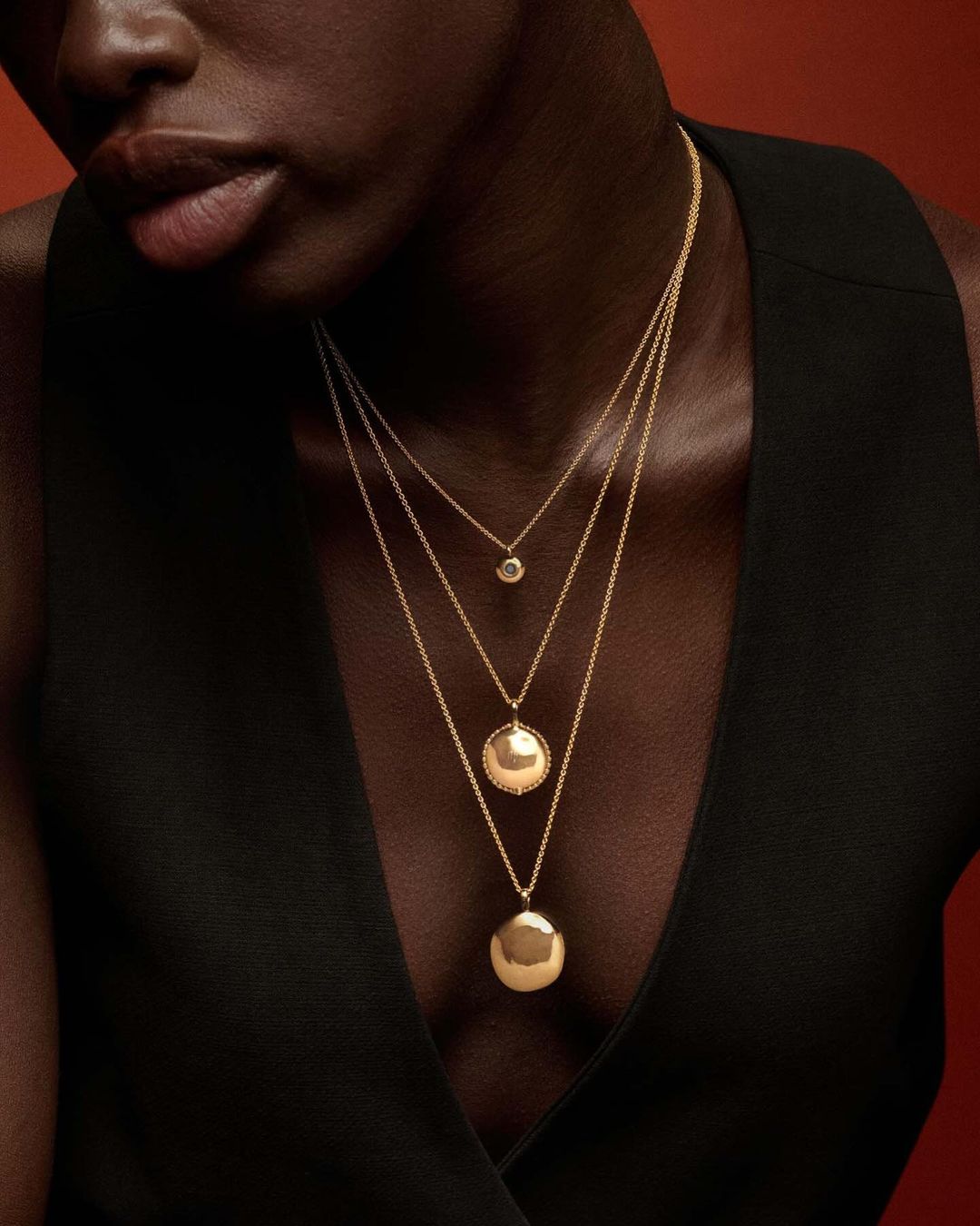 Close-up of a Black woman wearing three gold necklaces of varying lengths and pendants against a dark reddish-brown background.
