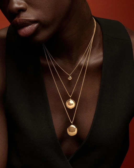 Close-up of a Black woman wearing three gold necklaces of varying lengths and pendants against a dark reddish-brown background.
