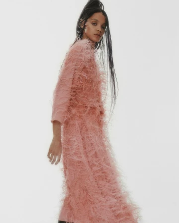 Model in a textured pink feathered dress with long dark braided hair standing against a light grey background.