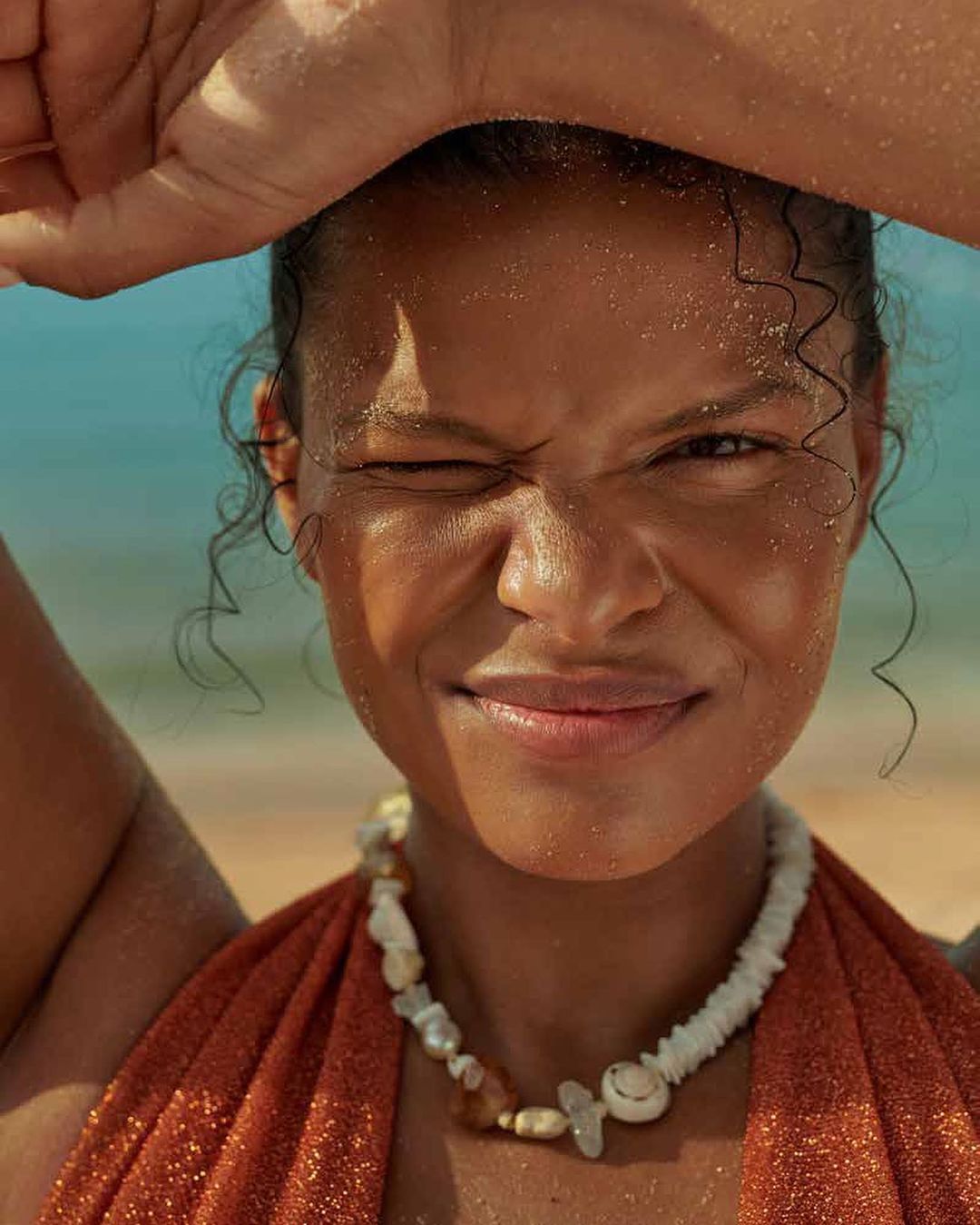 Close-up of a woman with dark skin and curly hair on a beach, wearing a shell necklace and orange top, shielding her eyes from the sun.