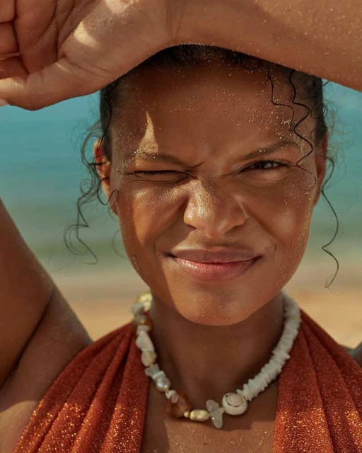 Close-up of a woman with dark skin and curly hair on a beach, wearing a shell necklace and orange top, shielding her eyes from the sun.