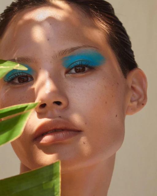 Close-up portrait of an adult female with dramatic teal eyeshadow and glossy hair, posing with a large green leaf.