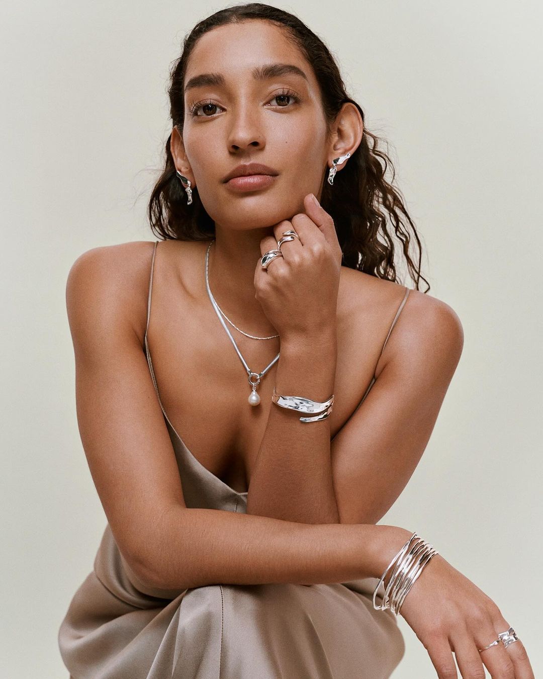 Portrait of a woman wearing silver jewelry including earrings, necklace, rings and bracelets against a light neutral background.