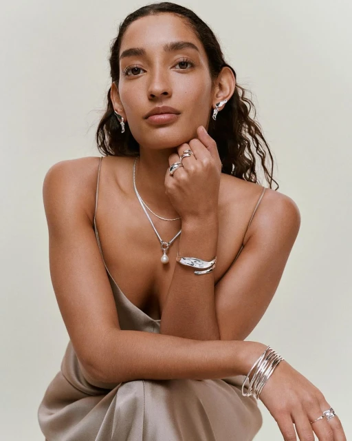 Portrait of a woman wearing silver jewelry including earrings, necklace, rings and bracelets against a light neutral background.