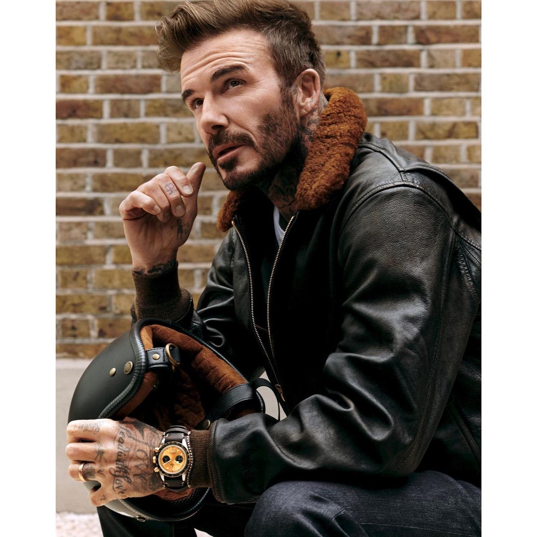 Portrait of adult male in leather jacket holding motorcycle helmet against a brick wall, wearing a wristwatch.