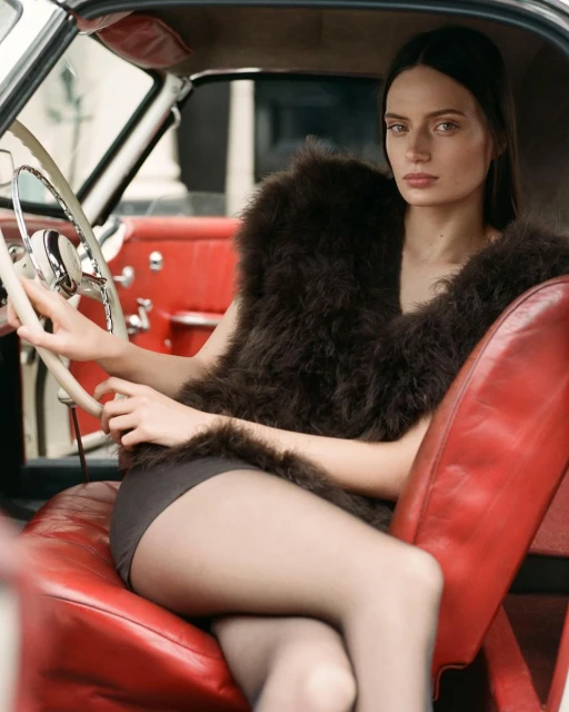 Elegant woman in a fur vest sits in the red leather interior of a vintage car, holding the steering wheel, in a fashion editorial.
