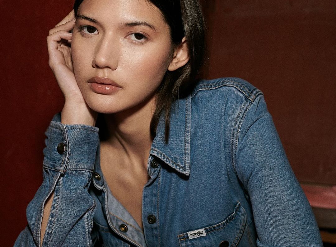 Close-up of a young woman wearing a denim jacket, posing with her head resting on her hand, against a dark red backdrop.