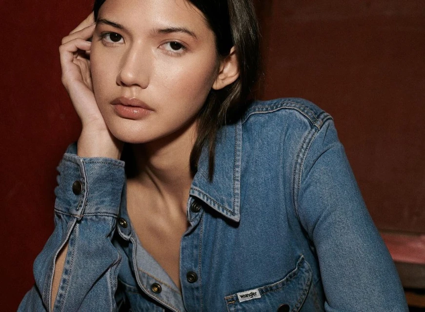 Close-up of a young woman wearing a denim jacket, posing with her head resting on her hand, against a dark red backdrop.