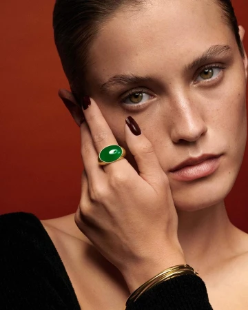 Close-up of a woman showcasing a green stone ring with gold band, complemented by gold bracelets, against a warm orange background.