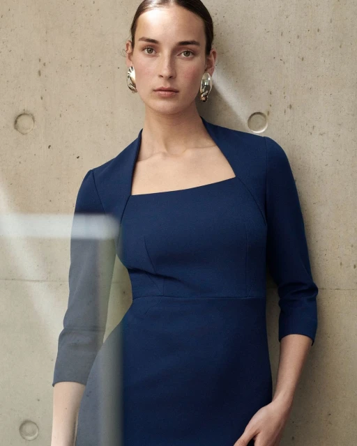Elegant adult woman in a blue dress and statement earrings posing against a textured concrete wall in a fashion editorial shot.