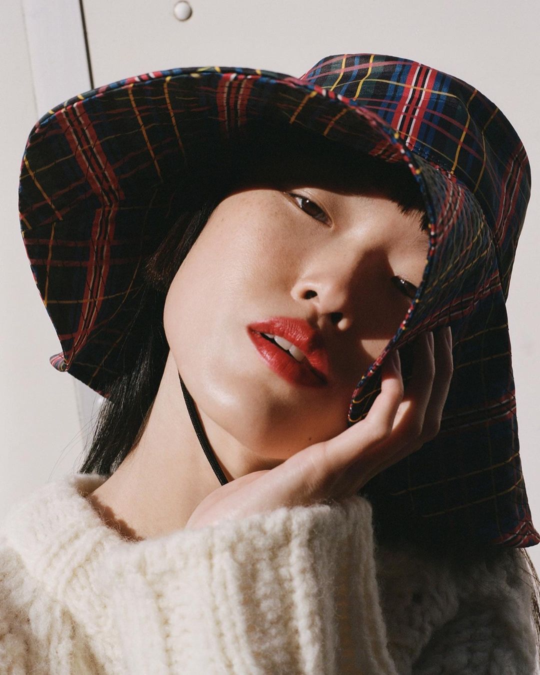 Close-up of a woman with dark hair and red lipstick, wearing a plaid bucket hat and a cream-colored sweater in soft studio light.