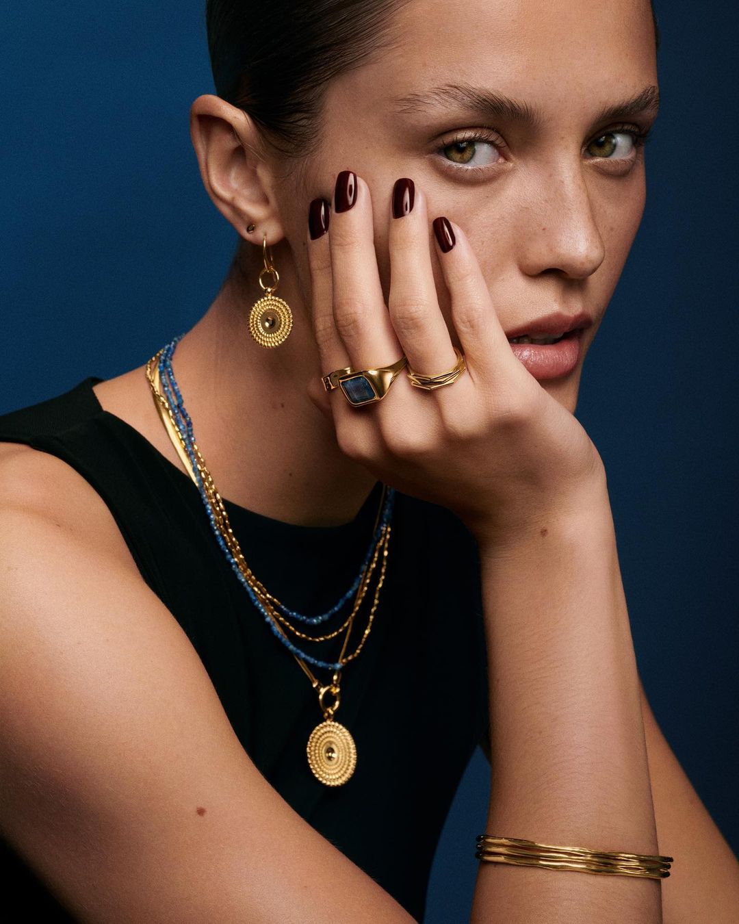 Close-up of adult woman with gold statement jewelry including necklace, earrings, rings, and bracelets on a dark blue background.