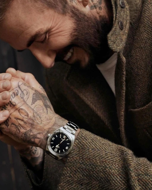 Close-up shot of David Beckham wearing a watch with a black face and silver wristband. He's wearing a herringbone jacket.