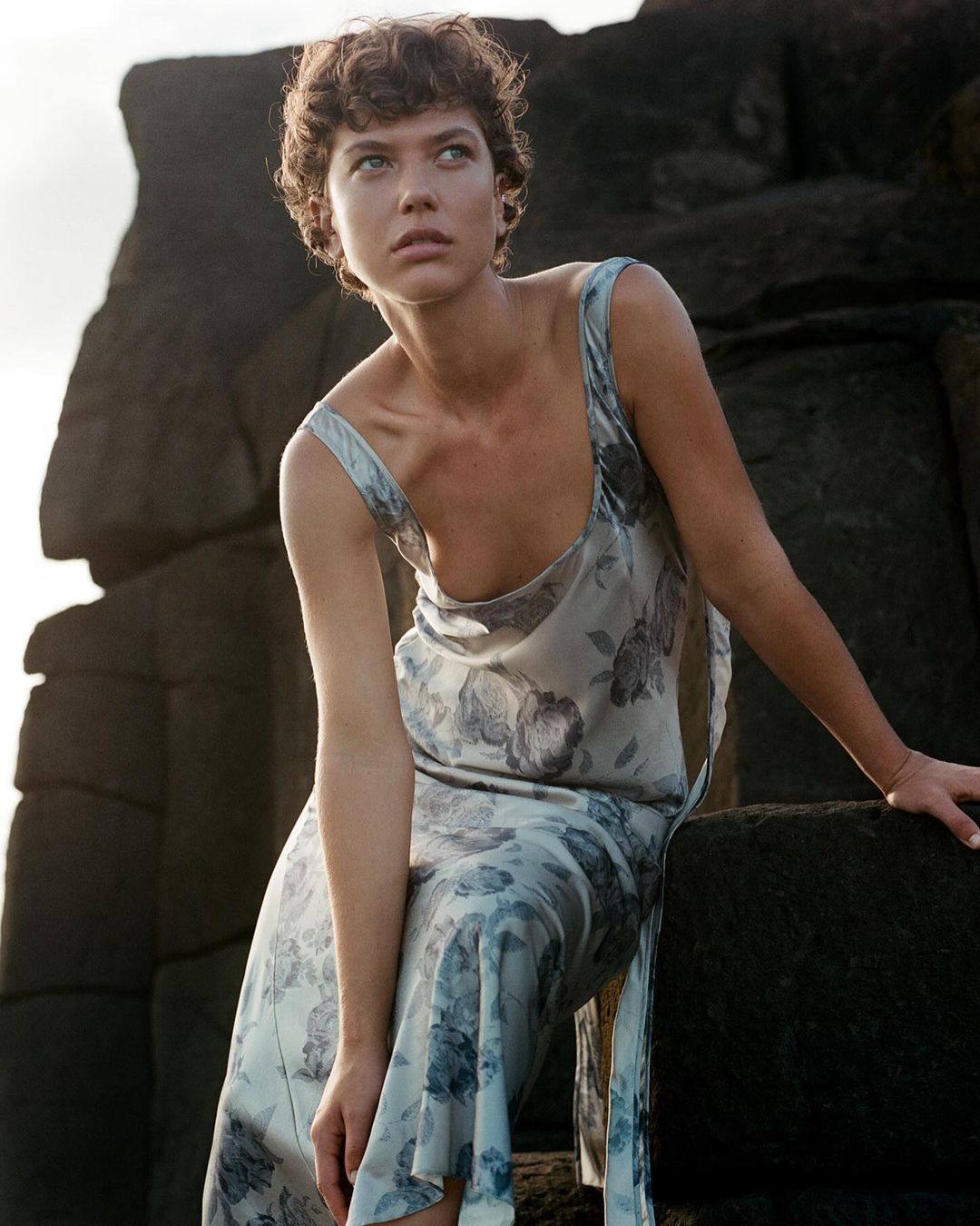 Woman in floral slip dress posing against rock formation. Portrait shot, outdoor fashion photography, blue and cream tones.