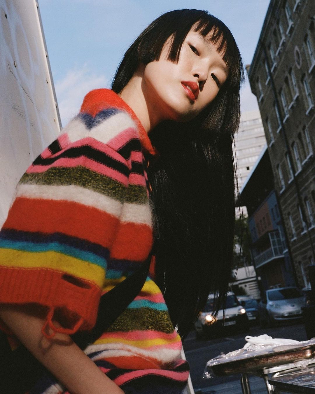 Young Asian woman with blunt bangs and striped sweater poses in an urban setting, with a building and parked cars visible in the background.