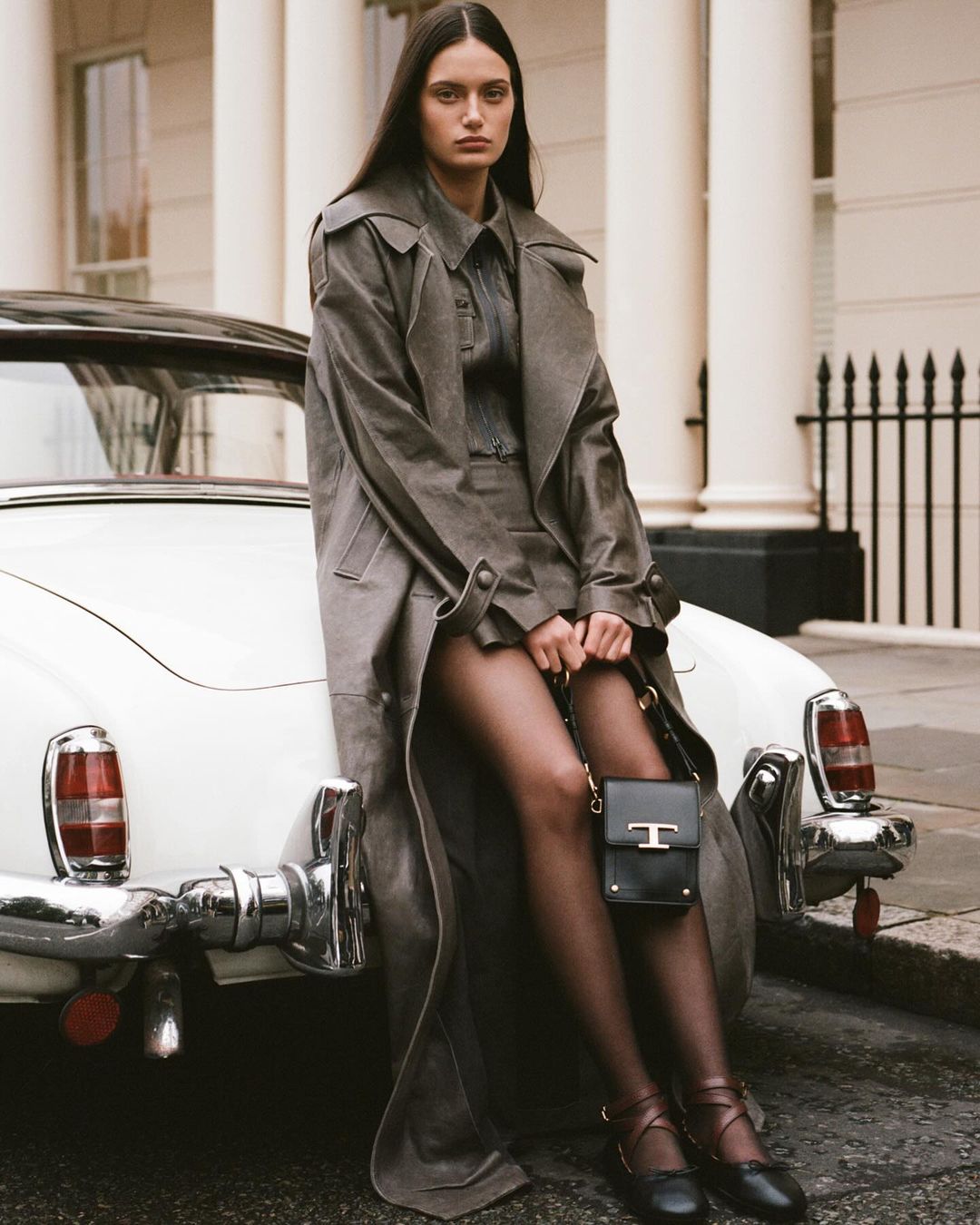 Model leaning against a vintage white car wearing a grey trench coat and holding a black handbag in London