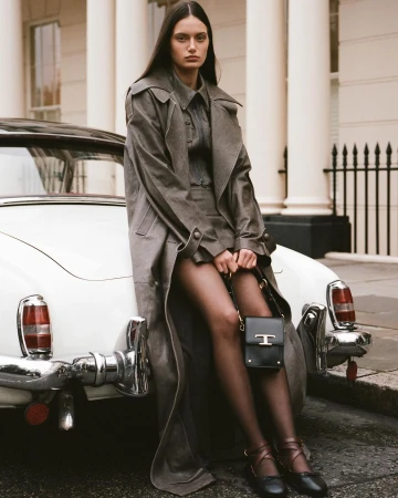 Model leaning against a vintage white car wearing a grey trench coat and holding a black handbag in London