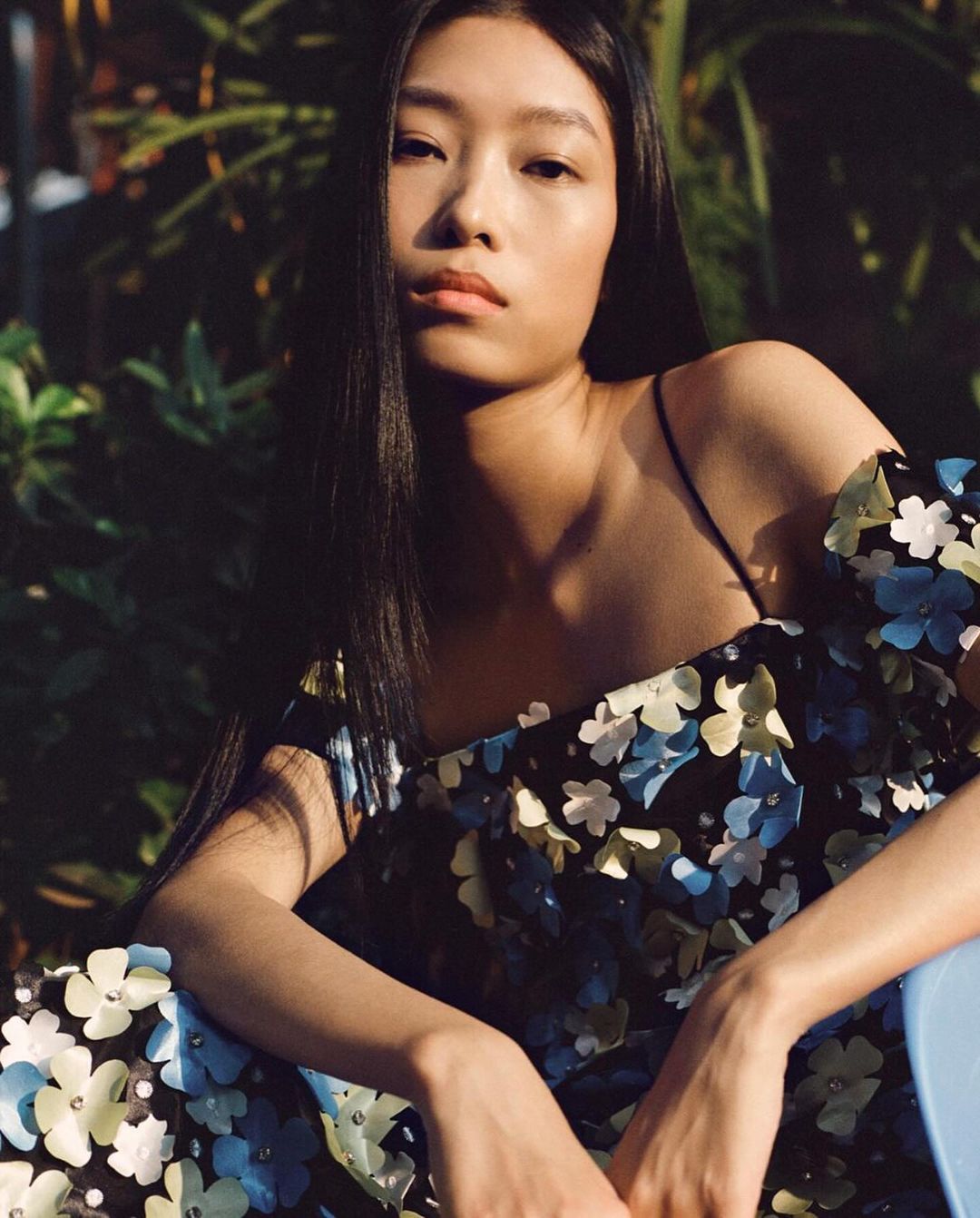 Portrait of a young Asian woman with long dark hair, wearing a floral dress, seated in natural light against a leafy green background.