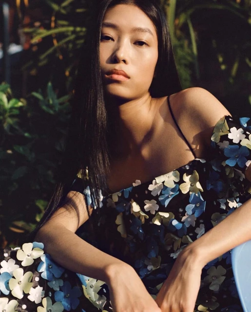 Portrait of a young Asian woman with long dark hair, wearing a floral dress, seated in natural light against a leafy green background.