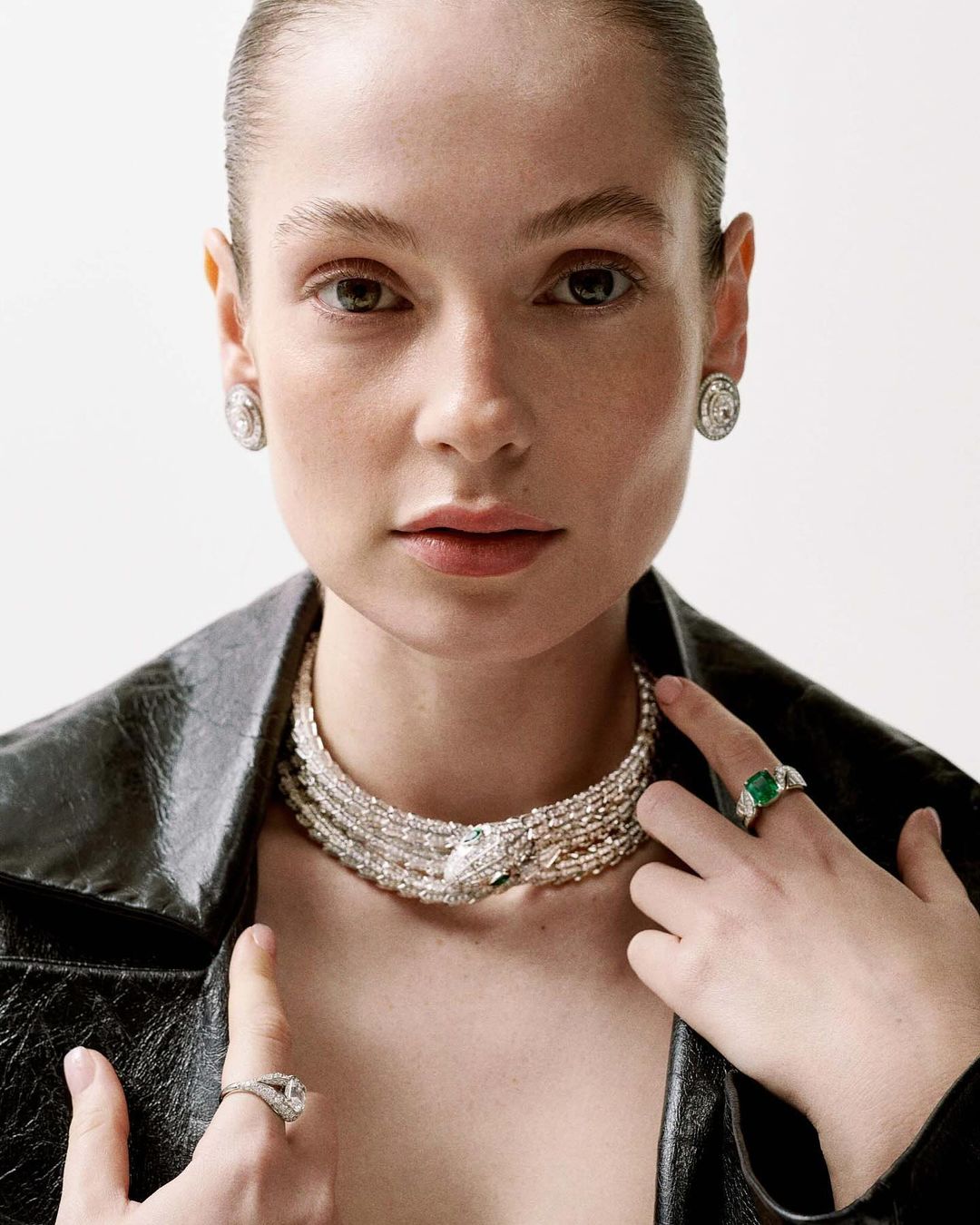 Close-up of a woman wearing a black jacket and jewelry including a multilayer choker necklace and rings, standing against a neutral background.