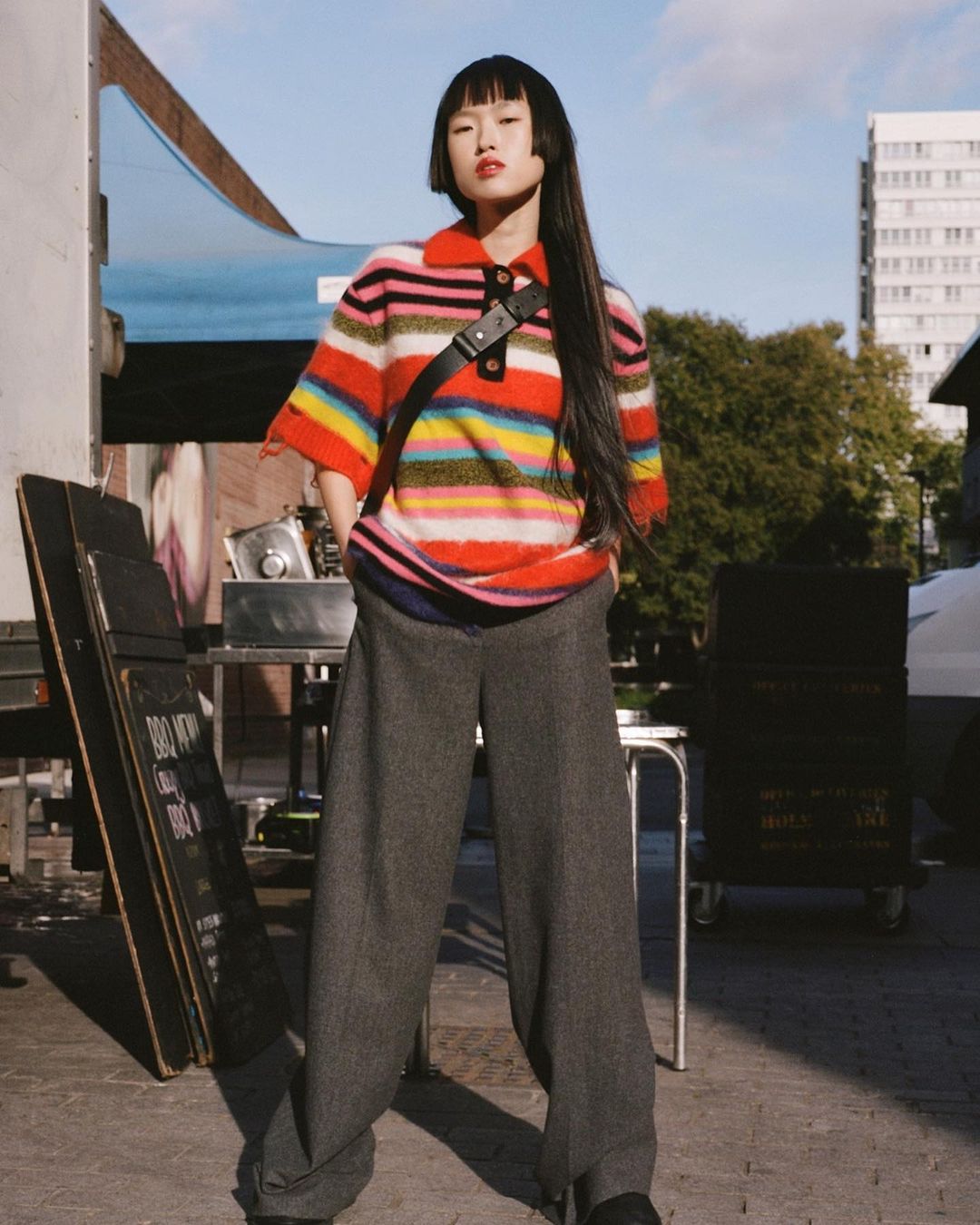Adult woman with long dark hair wears a colorful striped knit sweater, gray pants, and a black shoulder bag in an urban setting.