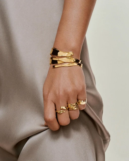 Close-up of a person's arm adorned with a gold cuff bracelet and several gold rings against a neutral dress background.
