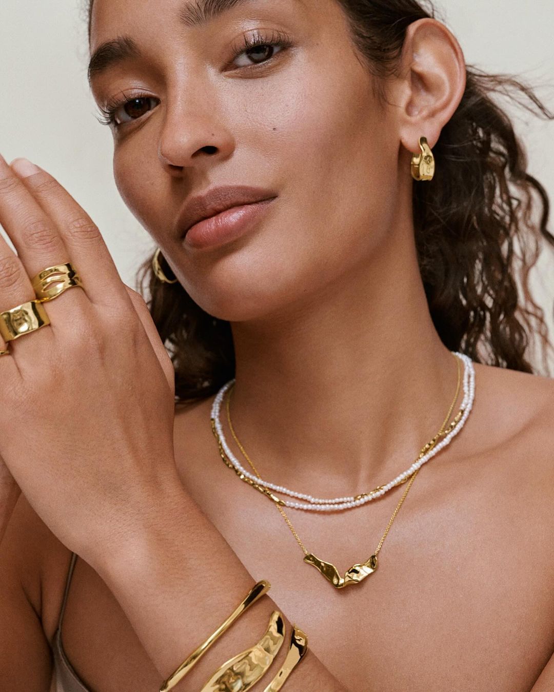 Close-up of an adult woman wearing gold jewelry, including rings, hoop earrings, necklace, and bracelets, against a light backdrop.