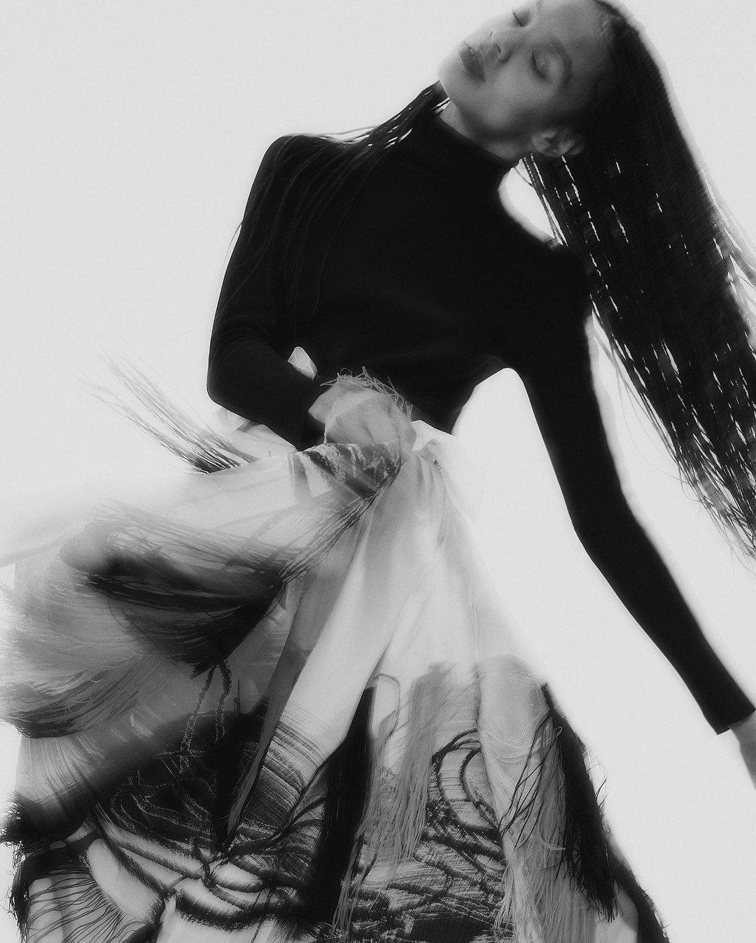 Monochrome image of an adult female model in a black turtleneck and flowing skirt with black patterns against a white backdrop.