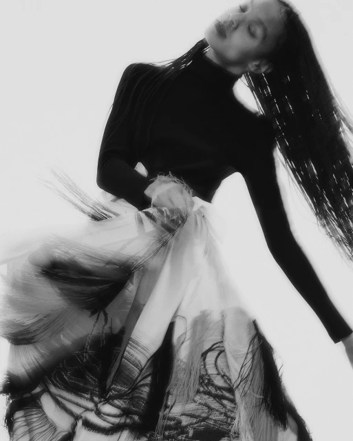 Monochrome image of an adult female model in a black turtleneck and flowing skirt with black patterns against a white backdrop.