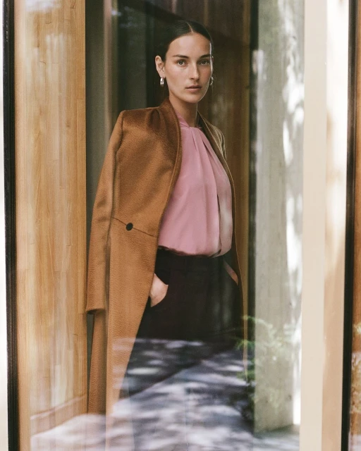 Adult female in a brown jacket and pink blouse stands indoors with natural lighting in a medium shot with selective focus.