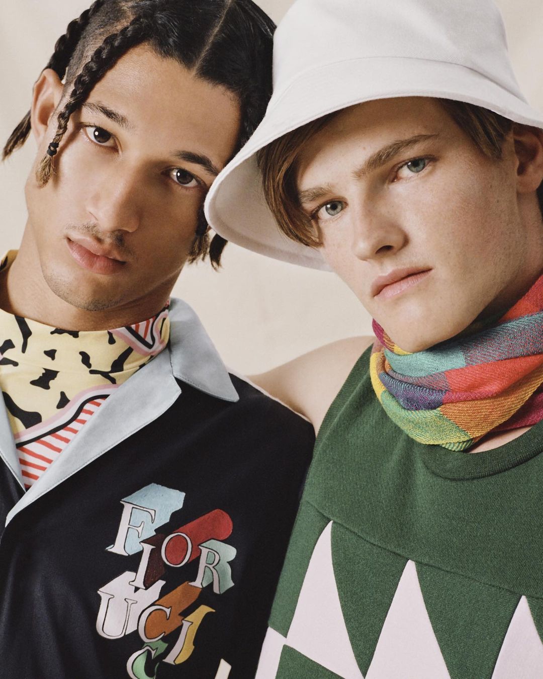 Two stylish men pose indoors wearing colorful tops and accessories in a fashion-forward studio portrait.