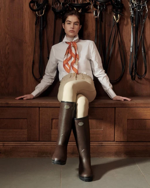 Woman in equestrian attire sitting on wooden bench in front of riding equipment inside a stable. Fashion, riding, sport theme.