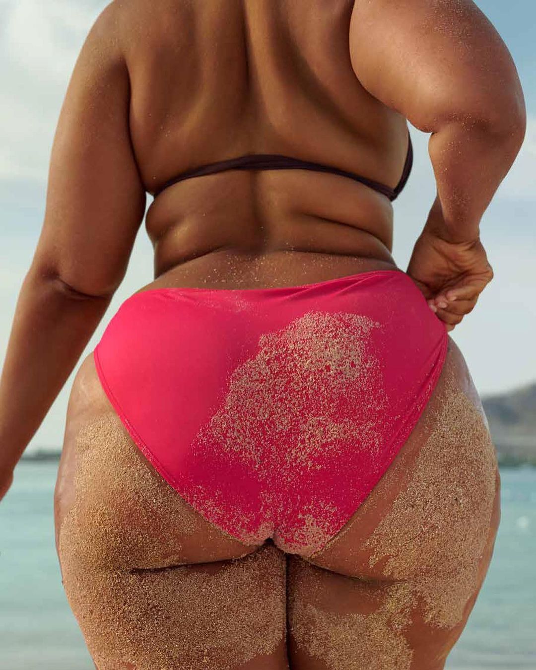Adult female stands on sandy beach with back to camera, wearing a pink bikini bottom, sandy skin, tropical water and sky.