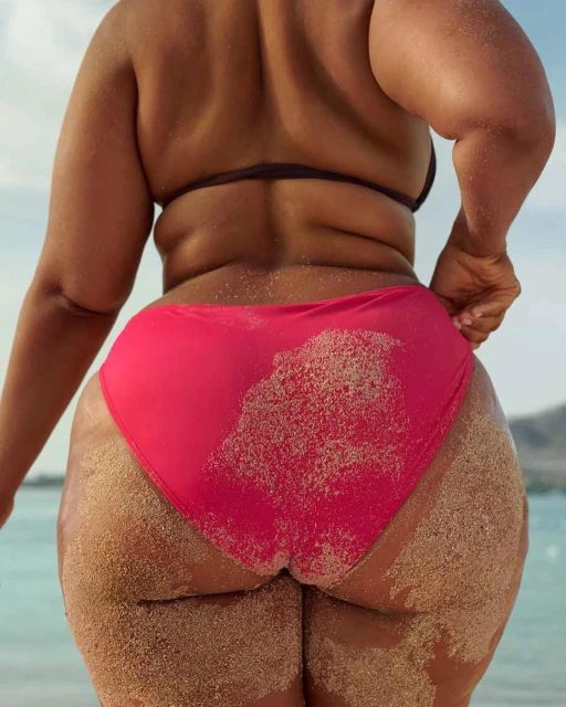 Adult female stands on sandy beach with back to camera, wearing a pink bikini bottom, sandy skin, tropical water and sky.