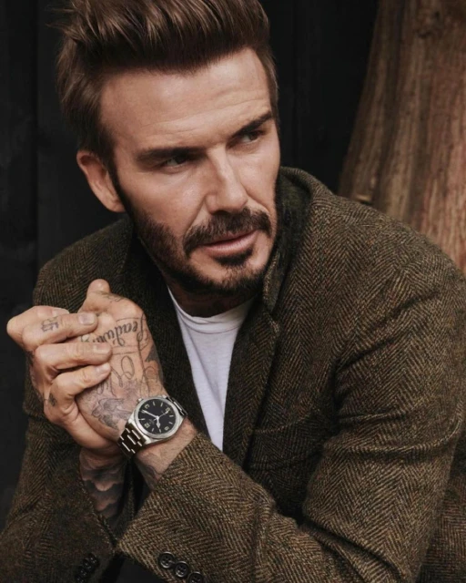 Close-up of adult male model David Beckham wearing a brown herringbone jacket and watch in an indoor setting with dark backdrop.