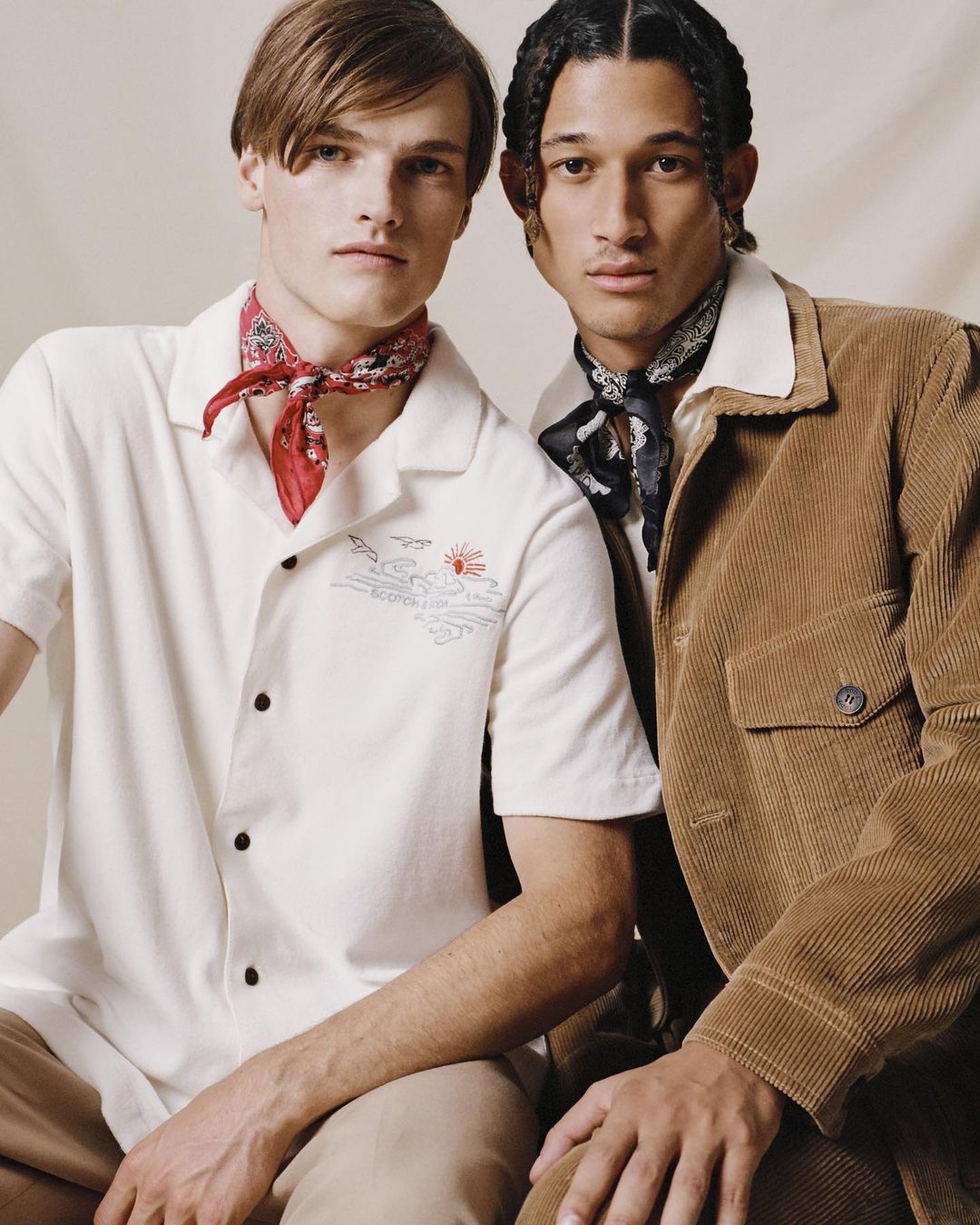 Two men pose indoors; one in a white shirt with an embroidered design and a red bandana, the other in a brown corduroy jacket and a black bandana.